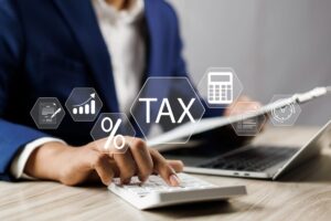 写真：書類を手に電卓を叩いているひとを背景に、「TAX」の文字やグラフ、電卓などのアイコンが浮かんでいる
