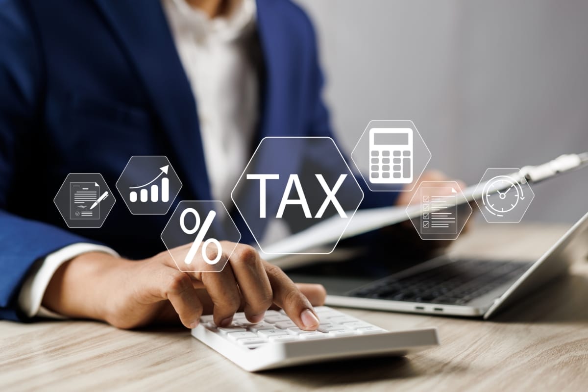 写真：書類を手に電卓を叩いているひとを背景に、「TAX」の文字やグラフ、電卓などのアイコンが浮かんでいる